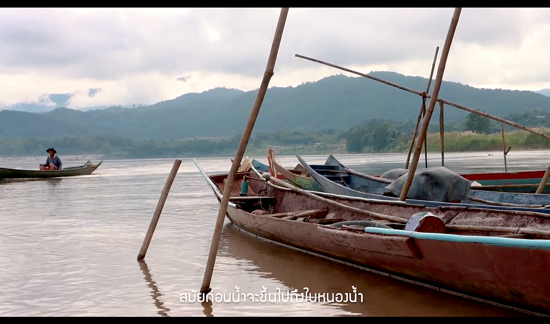Documentary still — wooden boats moored on the river with distant hills