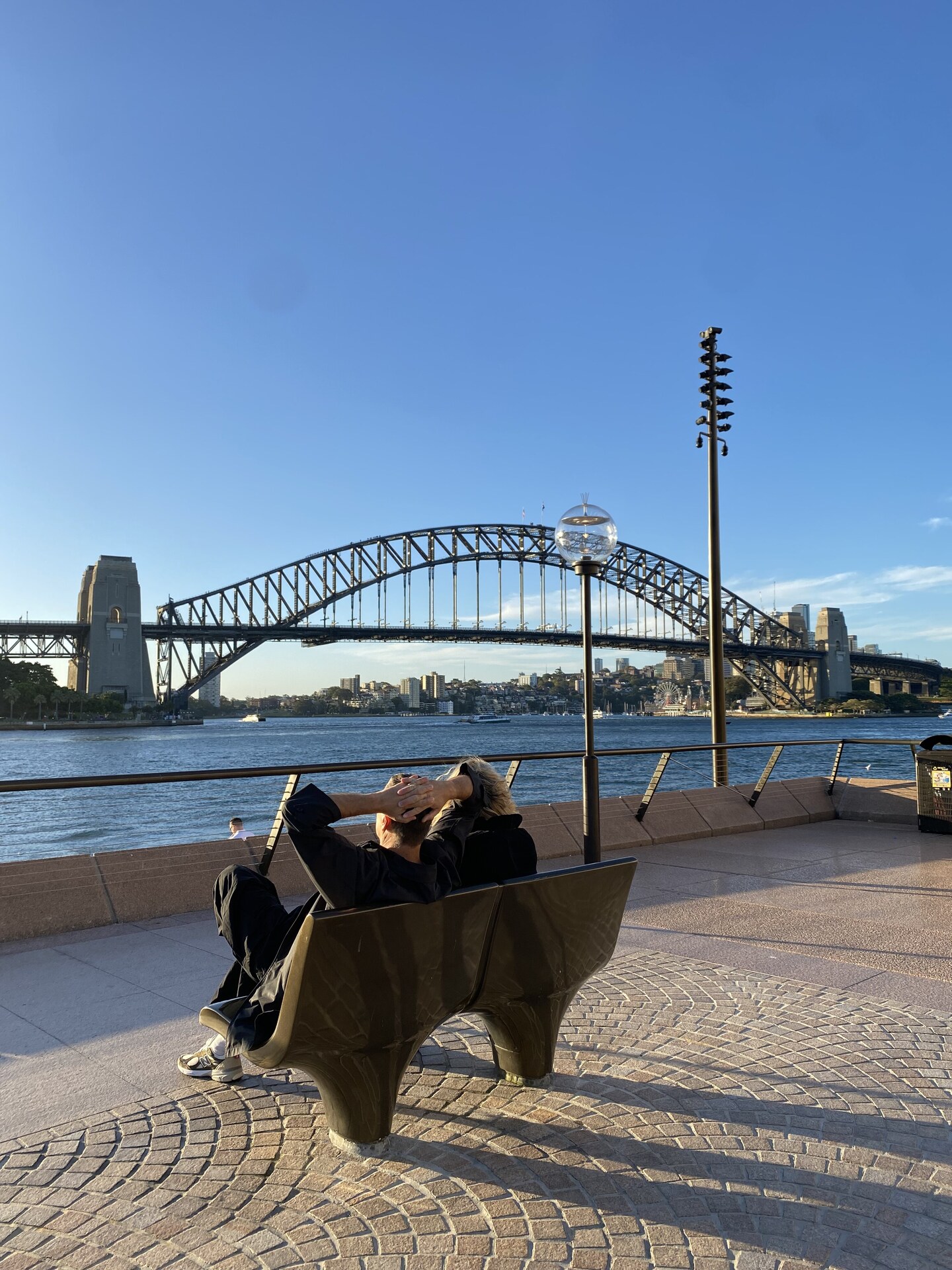 Two people sitting on a bench by the water with the Sydney Harbour Bridge behind them